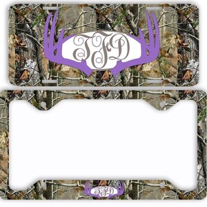 May include: A license plate and frame set featuring a camouflage pattern with a purple antler design. The antler shape frames a monogram in a decorative font. The license plate and frame are designed to fit a standard US vehicle.