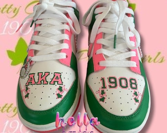 May include: Pair of white, green, and pink Nike trainers with white laces. The trainers feature the text "AKA" and "1908" in pink and green. The shoes have a green sole and pink accents.