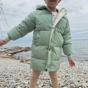 May include: A pale green hooded puffer jacket, worn over a cream sweater and tan leggings. The jacket features a zip closure and pockets. The person is wearing white trainers and standing on a pebble beach.