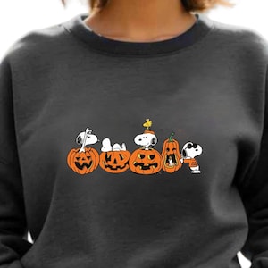 May include: Dark grey sweatshirt with a Halloween design. The design features Snoopy and Woodstock characters on carved pumpkins. The sweatshirt has a crew neck and long sleeves. The text "Belvity Embroidery" is visible in the upper left corner.