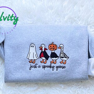 May include: A grey sweatshirt with a Halloween design. The design shows four cartoon geese dressed as a ghost, vampire, pumpkin, and skeleton. The text "just a spooky goose" is below the geese.