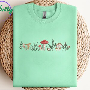 Może przedstawiać: Jasnozielony t-shirt z grafiką przedstawiającą grzyby i liście w odcieniach brązu, czerwieni i zieleni.
