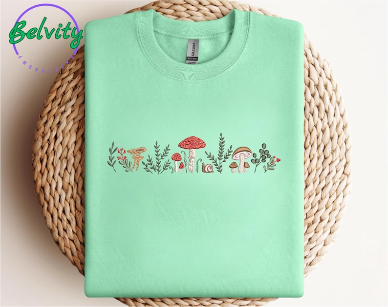 Może przedstawiać: Jasnozielony t-shirt z grafiką przedstawiającą grzyby i liście w odcieniach brązu, czerwieni i zieleni.