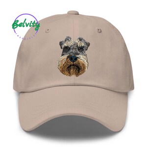 May include: A beige baseball cap featuring an embroidered portrait of a Schnauzer dog on the front panel. The dog has grey and black fur on its head with a tan-coloured beard and snout. The text Belvity is printed in green in the upper left corner.