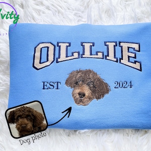 Peut inclure: Sweat-shirt bleu clair avec le nom "OLLIE" en blanc et bleu marine, et un portrait de chien marron. Le texte "EST 2024" est en dessous du nom. Une petite photo du chien est dans le coin inférieur gauche.