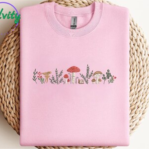 Może przedstawiać: R&oacute;żowy T-shirt z haftowaną linią grzyb&oacute;w i zieleni. Grzyby są w r&oacute;żnych kolorach, w tym czerwonym, brązowym i białym.