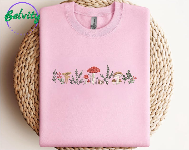 Może przedstawiać: R&oacute;żowy T-shirt z haftowaną linią grzyb&oacute;w i zieleni. Grzyby są w r&oacute;żnych kolorach, w tym czerwonym, brązowym i białym.