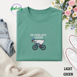 May include: A light green t-shirt featuring an embroidered graphic of a white goose wearing a blue helmet while riding a blue bicycle. The text above the goose reads, "ON YOUR LEFT, LOSERS" in white capital letters. The shirt has a crew neckline.