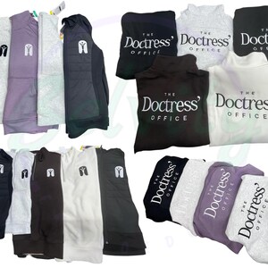 May include: A collection of various sweatshirts and hoodies in different colors, including gray, black, white, and purple. Some of the sweatshirts have the text "The Doctress' Office" printed on them.