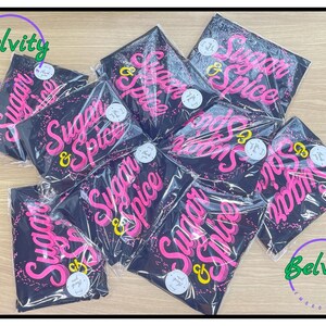 May include: A stack of black t-shirts with the words "Sugar & Spice" printed in pink on each shirt. The shirts are packaged in plastic bags and have a Belvity logo on the top and bottom of the image.