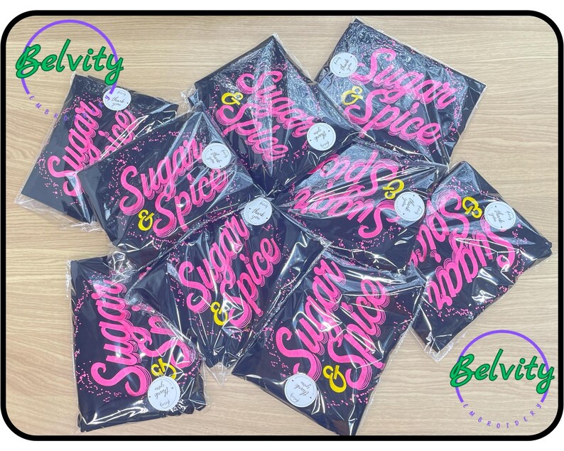 May include: A stack of black t-shirts with the words "Sugar & Spice" printed in pink on each shirt. The shirts are packaged in plastic bags and have a Belvity logo on the top and bottom of the image.