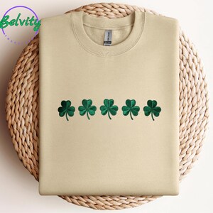 May include: A beige sweatshirt featuring five embroidered green shamrocks. The garment has a crew neck and long sleeves. The brand name "Belvity" is in a circle in the upper left corner.