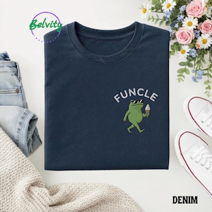 May include: A navy blue t-shirt featuring an embroidered graphic of a green frog wearing black sunglasses and holding a white ice cream cone. The word FUNCLE is embroidered in white capital letters in an arc above the frog character.