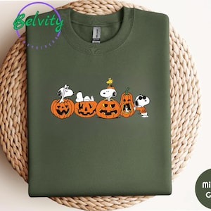 May include: Military green sweatshirt featuring Snoopy and friends with carved orange pumpkins. The graphic includes Snoopy characters in Halloween costumes. The sweatshirt has a crew neck and long sleeves.