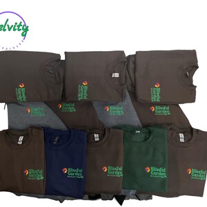 May include: A stack of brown t-shirts with the text "Blissful Garden" embroidered on the front. The shirts are folded and stacked on top of each other.