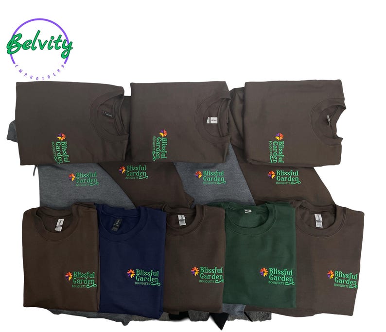 May include: A stack of brown t-shirts with the text "Blissful Garden" embroidered on the front. The shirts are folded and stacked on top of each other.