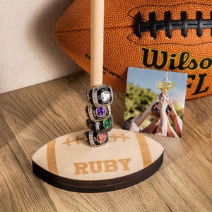 May include: A wooden football-shaped display stand with the word "RUBY" engraved on it. A stack of championship rings with colorful gemstones sits on a wooden dowel. A football and a photo of a trophy are in the background.