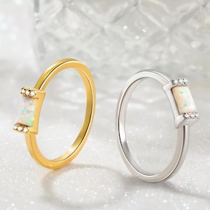 May include: Two simple rings with a rectangular opal stone set in a bezel setting. The ring on the left is gold and the ring on the right is silver.