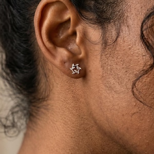 May include: A close-up of a person's ear with a silver stud earring. The earring is a small, circular design with a floral or leaf-like pattern. The skin tone is medium, and the background is blurred.