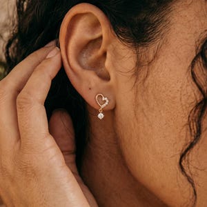 May include: A gold-colored earring with a heart-shaped design and a dangling diamond-like stone. The earring is worn on the ear of a person with dark curly hair. The earring is a delicate piece of jewelry.