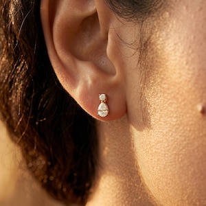 May include: A gold earring featuring two clear gemstones. The top gemstone is round, and the bottom is a pear shape. The earring is worn on an ear. The person has dark hair.