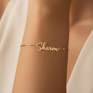 May include: A gold-colored bracelet with the name "Sharon" in cursive script. The delicate chain bracelet is worn on a wrist, with the name centered. The background is a soft, neutral color.