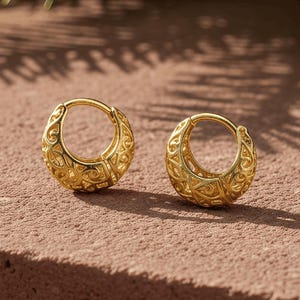 May include: A pair of gold-colored hoop earrings with an intricate, embossed design. The earrings are displayed on a reddish-brown surface, with evergreen branches in the background. The earrings have a diameter of approximately 0.75 inches.