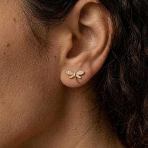 以下が含まれることがあります： 金色のトンボのスタッドピアス。羽には小さな透明な宝石がちりばめられています。ピアスは耳に装着され、繊細なデザインと輝くディテールを際立たせています。ジュエリーです。