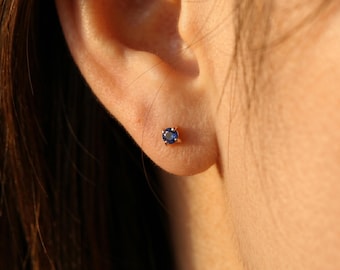 May include: A close-up of a small, round, dark blue gemstone stud earring set in a gold-colored metal. The earring is worn on an ear, showcasing the jewelry's size and design. The earring is a classic and elegant piece.