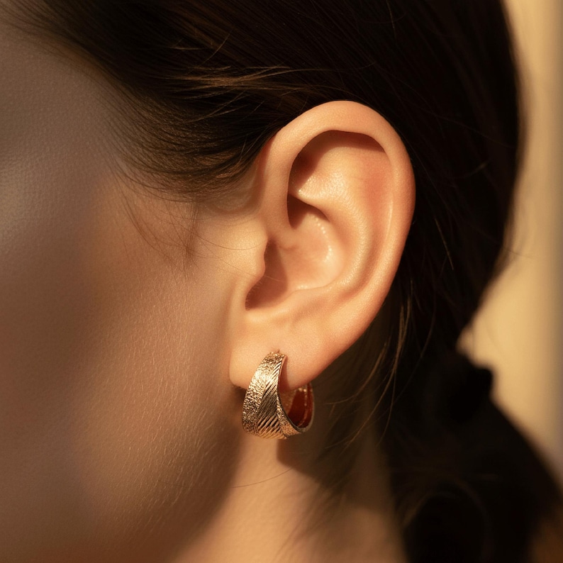 May include: A close-up of a person's ear wearing gold-coloured hoop earrings. The earrings have a textured surface and a curved shape. The background is blurred, with warm lighting.