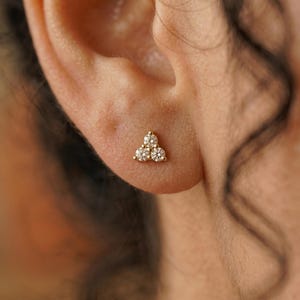 May include: A gold stud earring with three clear, round gemstones arranged in a triangular cluster. The earring is set against the backdrop of a person's ear, highlighting the jewelry's delicate design and sparkle.