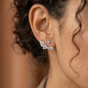 May include: A person's ear is shown with two earrings. One is a small, square stud, and the other is a cluster of white gemstones in a curved design. The earrings are set against the skin, highlighting their sparkle and intricate design.