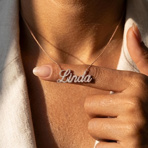May include: A rose gold-toned necklace featuring a cursive nameplate pendant that spells the name Linda. The pendant is encrusted with small, clear, sparkling stones throughout the letters, suspended from a delicate, matching rose gold-toned chain.