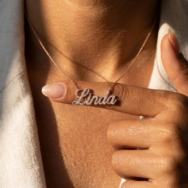 May include: A rose gold-toned necklace featuring a cursive nameplate pendant that spells the name Linda. The pendant is encrusted with small, clear, sparkling stones throughout the letters, suspended from a delicate, matching rose gold-toned chain.