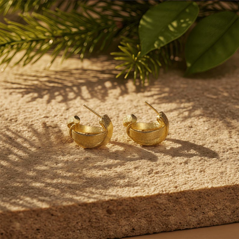 May include: A pair of gold hoop earrings with a textured finish. The earrings are presented on a light-coloured, textured surface, with green foliage in the background. The earrings have a distinctive design.
