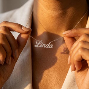 May include: A silver-toned name necklace featuring the name Linda in a cursive script font. The letters are encrusted with small, clear, sparkling stones. The pendant is attached to a delicate, thin metal chain that rests around the neck area.