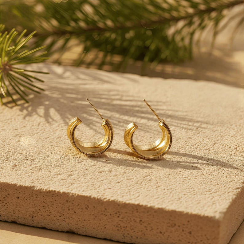 May include: A pair of gold hoop earrings. Each earring has a thick, rounded design with a textured surface. The earrings are displayed on a light-coloured, textured surface, with a blurred green plant in the background.