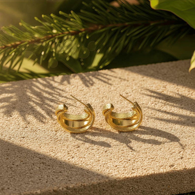 May include: A pair of gold hoop earrings. The earrings feature a textured outer band and a smooth inner band. They are presented on a light-coloured, textured surface, with green foliage in the background.