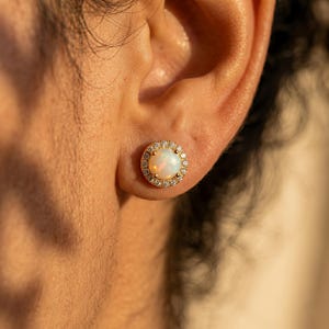 May include: A gold stud earring with a round, white opal center stone, surrounded by a halo of small, clear crystals. The earring is set in a gold metal setting and is worn on the ear.