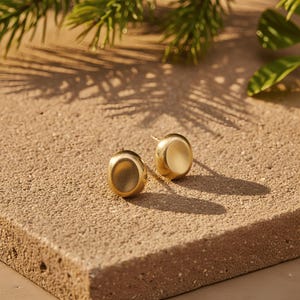 May include: Two gold-colored stud earrings with a rounded, oval design. The earrings are set on a textured, light brown surface, with shadows and greenery in the background. The earrings have a matte finish and a simple, elegant style.