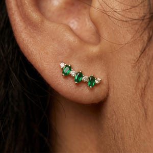 May include: A gold ear climber earring with three oval emeralds and small white gemstones. The emeralds are a vivid green, and the white gemstones add sparkle. The earring is designed to curve along the ear.