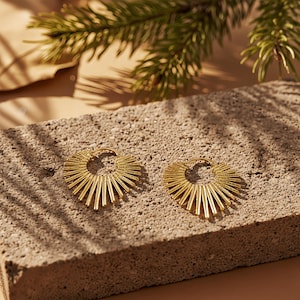 Könnte beinhalten: Zwei goldfarbene Creolen mit Sonnenstrahl-Design, die auf einer strukturierten grauen Oberfläche liegen. Die Ohrringe haben eine kreisförmige Form mit radialen Linien, die einen Sternen-Effekt erzeugen. Ein Zweig mit grünen Blättern ist im Hintergrund zu sehen.