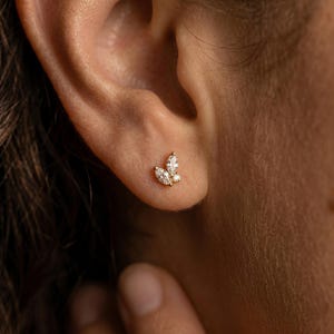 May include: A gold stud earring featuring three clear, marquise-cut gemstones arranged in a leaf-like design. The earring is set in a gold metal setting and is worn on the ear.
