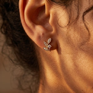 May include: A gold-colored leaf-shaped stud earring with small, clear gemstones. The earring has a central square gemstone and three leaf-like structures with smaller gemstones. The earring is worn on an ear.