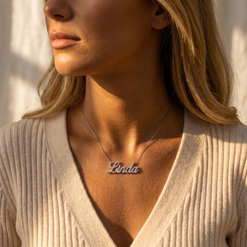 May include: A silver-toned metal nameplate necklace featuring the name Linda in a cursive script font. The pendant is suspended from a delicate, thin chain that rests against the collarbone, worn with a beige ribbed knit top.