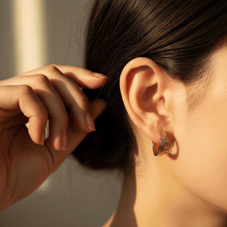 May include: Close-up of a person's ear with a pair of gold hoop earrings. The earrings have a textured surface and are a warm, golden colour. The person's hand is touching their hair.