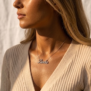 May include: A silver-toned nameplate necklace featuring the name Linda in a cursive script font. The letters are encrusted with small, clear, sparkling stones. The pendant is attached to a delicate, thin metal chain that rests against the collarbone.