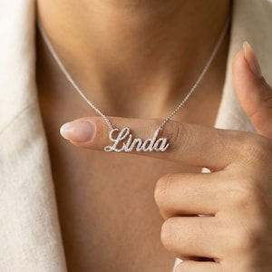 Op de afbeelding: Een zilveren ketting met de naam "Linda" in sierlijke letters. De hanger is bezet met kleine, glinsterende steentjes. De ketting wordt getoond tegen een neutrale achtergrond.