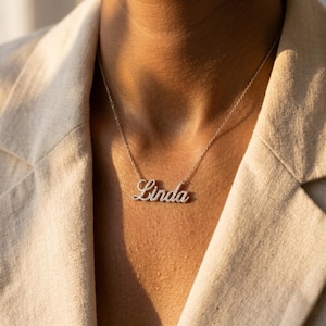 May include: A silver-toned necklace featuring a cursive nameplate pendant that reads Linda. The pendant is encrusted with small, clear, sparkling stones and is suspended from a delicate, thin silver-toned chain that rests against the collarbone.
