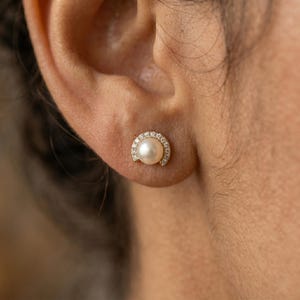 May include: A gold stud earring with a central white pearl, encircled by a halo of small, clear gemstones. The earring is set against the backdrop of a person's ear and shoulder.